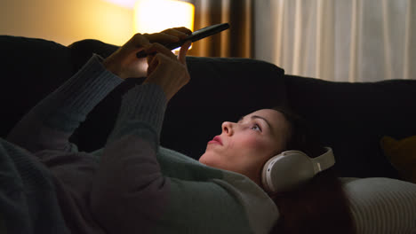 Woman-Wearing-Wireless-Headphones-Lying-On-Sofa-At-Home-At-Night-Streaming-Or-Looking-At-Online-Content-On-Mobile-Phone